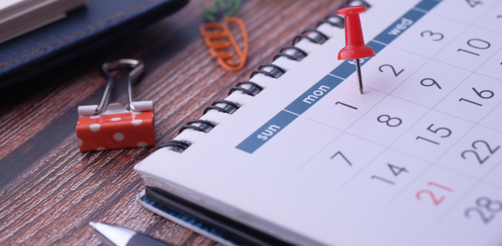 Close-up of a calendar with a red pushpin marking the 1st day, surrounded by a polka-dotted binder clip and stationery items on a wooden desk