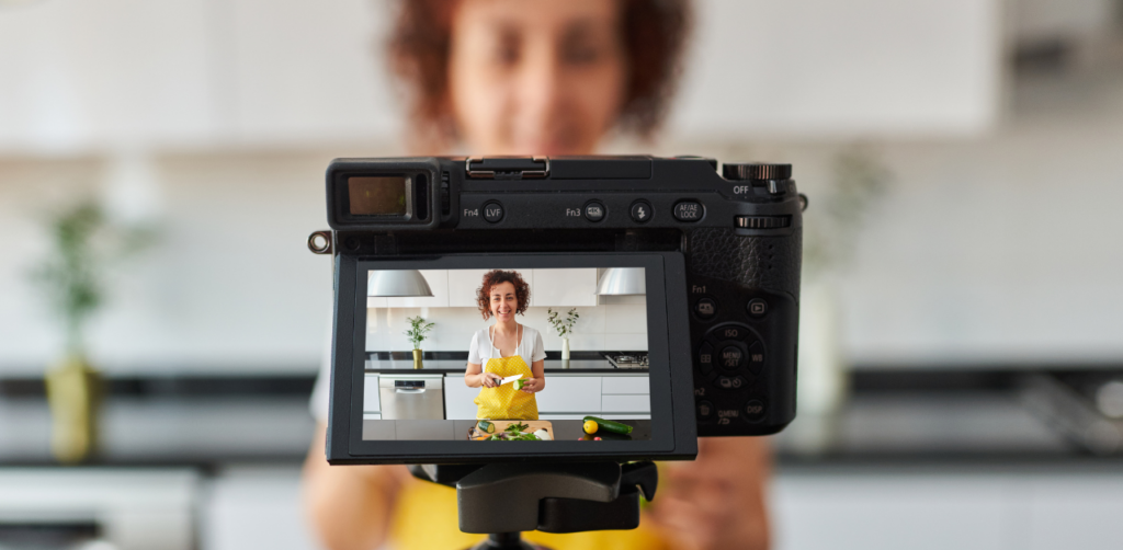 Digital camera recording a woman in a kitchen as she prepares food while wearing a yellow apron, with the scene visible on the camera's display