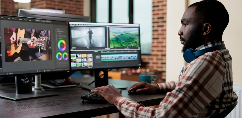 Man wearing headphones around his neck works on video editing at a dual-monitor setup, showing footage of a guitarist and nature scenes. A cup sits on the desk