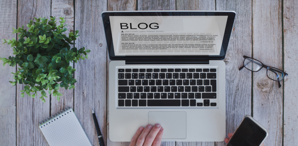 Overhead view of a laptop displaying a blog page, surrounded by a notepad, pen, glasses, a smartphone, and a potted plant on a wooden table