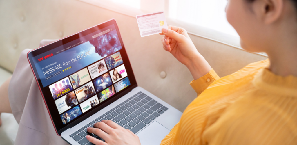 Person holding a credit card while browsing a video streaming platform on a laptop, showing movie and TV show thumbnails