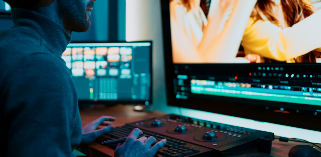 Person sitting at a desk, editing video content on a large monitor with editing software open. Bright screens and a control panel with knobs and sliders are visible