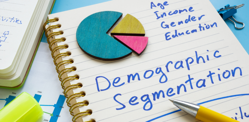 Spiral notebook with the words Demographic Segmentation written in blue, surrounded by a pie chart and a yellow pen