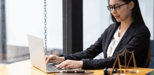 A businesswoman in glasses types on a laptop at a wooden desk with a gavel and legal scale beside her