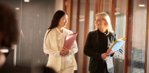 Two professionally dressed women walking and smiling, holding folders, in a well-lit office hallway with large windows