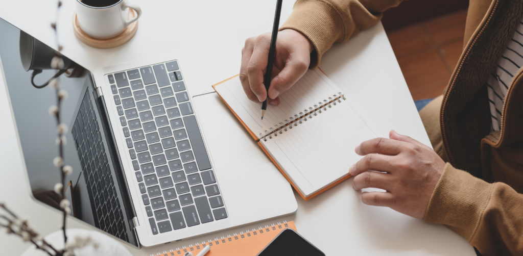 Person taking notes beside a laptop, symbolizing planning and strategy development in outreach marketing campaigns