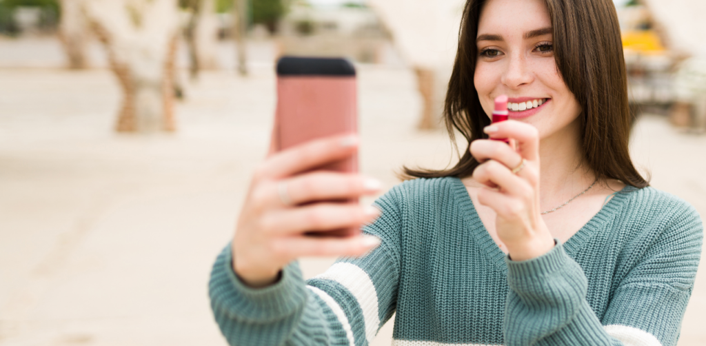 Woman applying lipstick while filming on smartphone, representing beauty influencer content creation