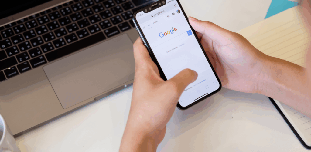 Person holding smartphone with Google homepage open next to a laptop