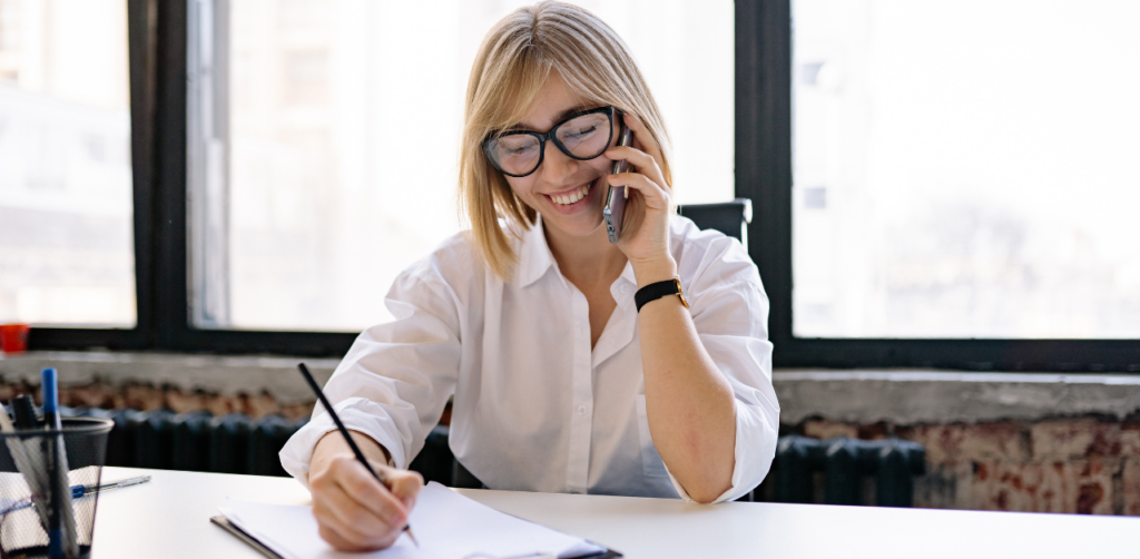 Smiling businesswoman talking on the phone while writing notes at her desk in a bright modern office
